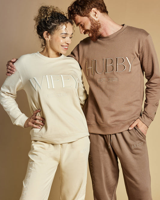 Man and woman wearing matching 'WIFE' and 'HUBBY' sweatshirt sets on a beige background