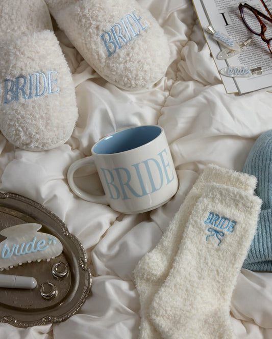 A styled flat-lay of bridal-themed home accessories on white satin bedding, including fluffy white slippers and socks with light blue embroidered 'BRIDE' text, a white mug with a light blue interior and large 'BRIDE' text, a blue knit beanie, reading glasses, and a small decorative tray.