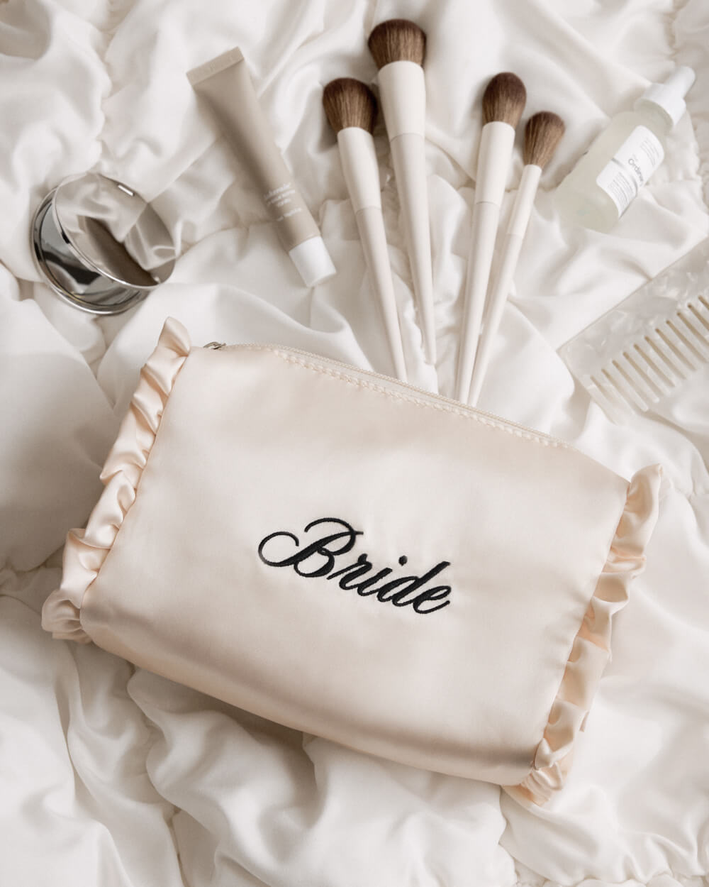 A cream satin makeup bag with ruffled trim and the word 'Bride' embroidered in black script, styled on white satin fabric with makeup brushes, a tube, a compact mirror, and a comb.