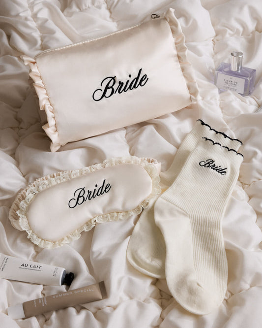 A coordinated bridal gift set arranged on white satin bedding, featuring a cream pillow with ruffles and the word 'Bride' in black script, a matching eye mask with ruffles and 'Bride', a cream sock with black trim and 'Bride' text, a small perfume bottle, a tube labeled 'AU LAIT', and a beige tube labeled 'SUMMER SPECIAL'.