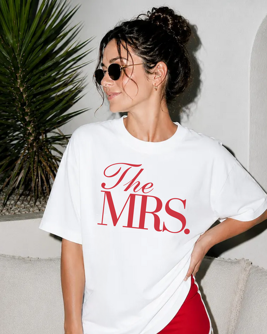 A woman wearing a white graphic t-shirt with 'The MRS.' printed in red, styled with red shorts and sunglasses, posing on a light-colored sofa next to a green plant.