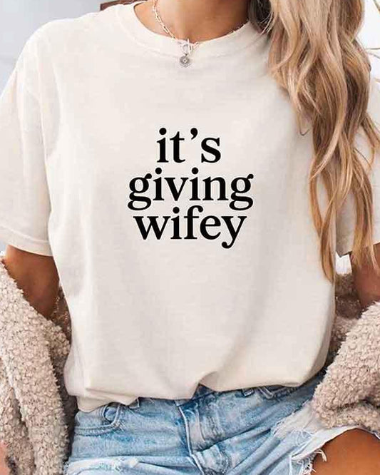 Woman wearing a white t-shirt with 'it's giving wifey' text, sitting on a couch.