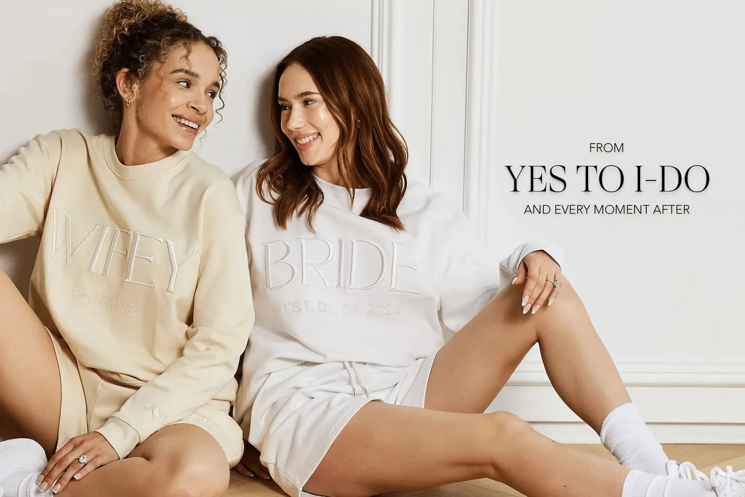 Two women sitting side by side wearing matching white and beige sweatshirt and shorts lounge sets with embossed text reading 'WIFEY' and 'BRIDE' respectively, in a bright indoor setting with text on the right reading 'FROM YES TO I-DO AND EVERY MOMENT AFTER'.