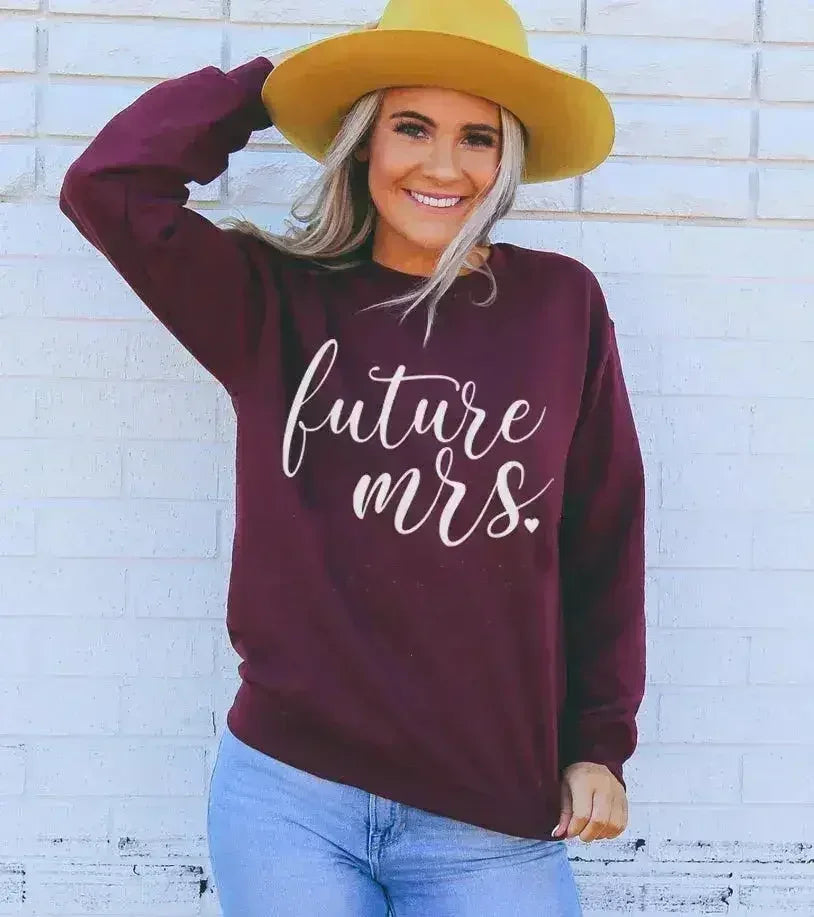 Woman wearing a maroon sweatshirt with 'future mrs.' text against a white brick wall.