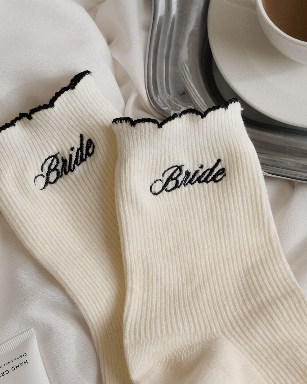 White socks with 'Bride' embroidery on a white surface with a cup of coffee in the background.
