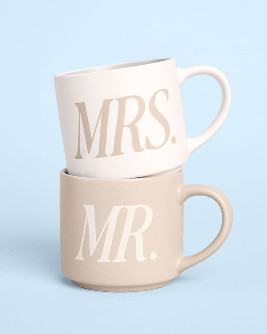 Two ceramic mugs stacked on top of each other against a light blue background. The top mug is white with the word 'MRS.