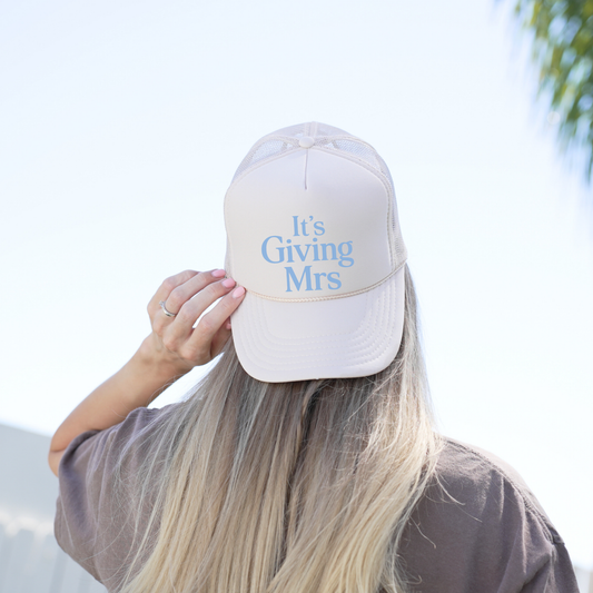 Person wearing a white cap with 'It's Giving Mrs.' text against a blurred outdoor background
